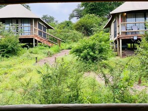 Two wooden buildings with balconies, surrounded by dense greenery and a dirt path.