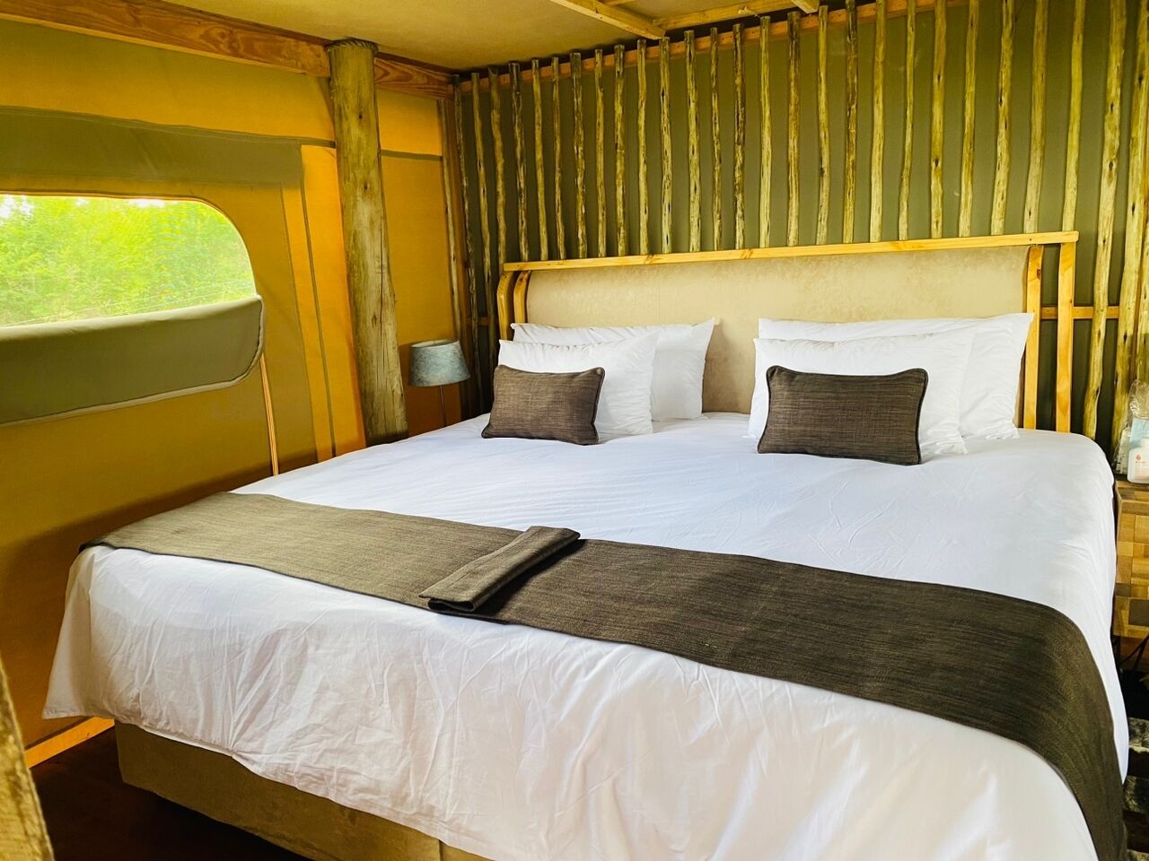 A bed with white bedding and a brown throw, a wooden headboard, and a window with a view of greenery.