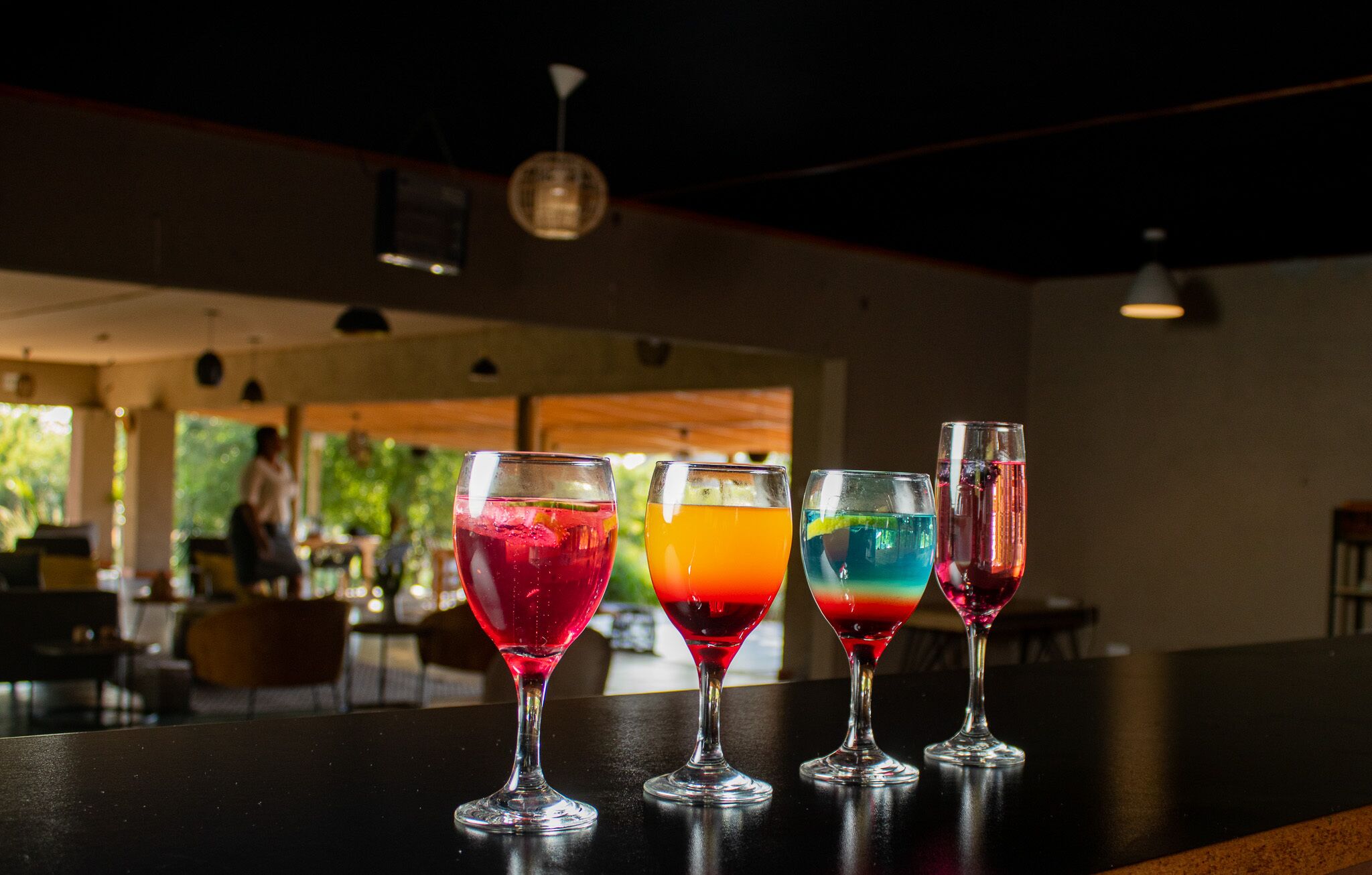 Four colorful cocktails on a bar counter in a restaurant with a modern interior and a view of the outdoors.