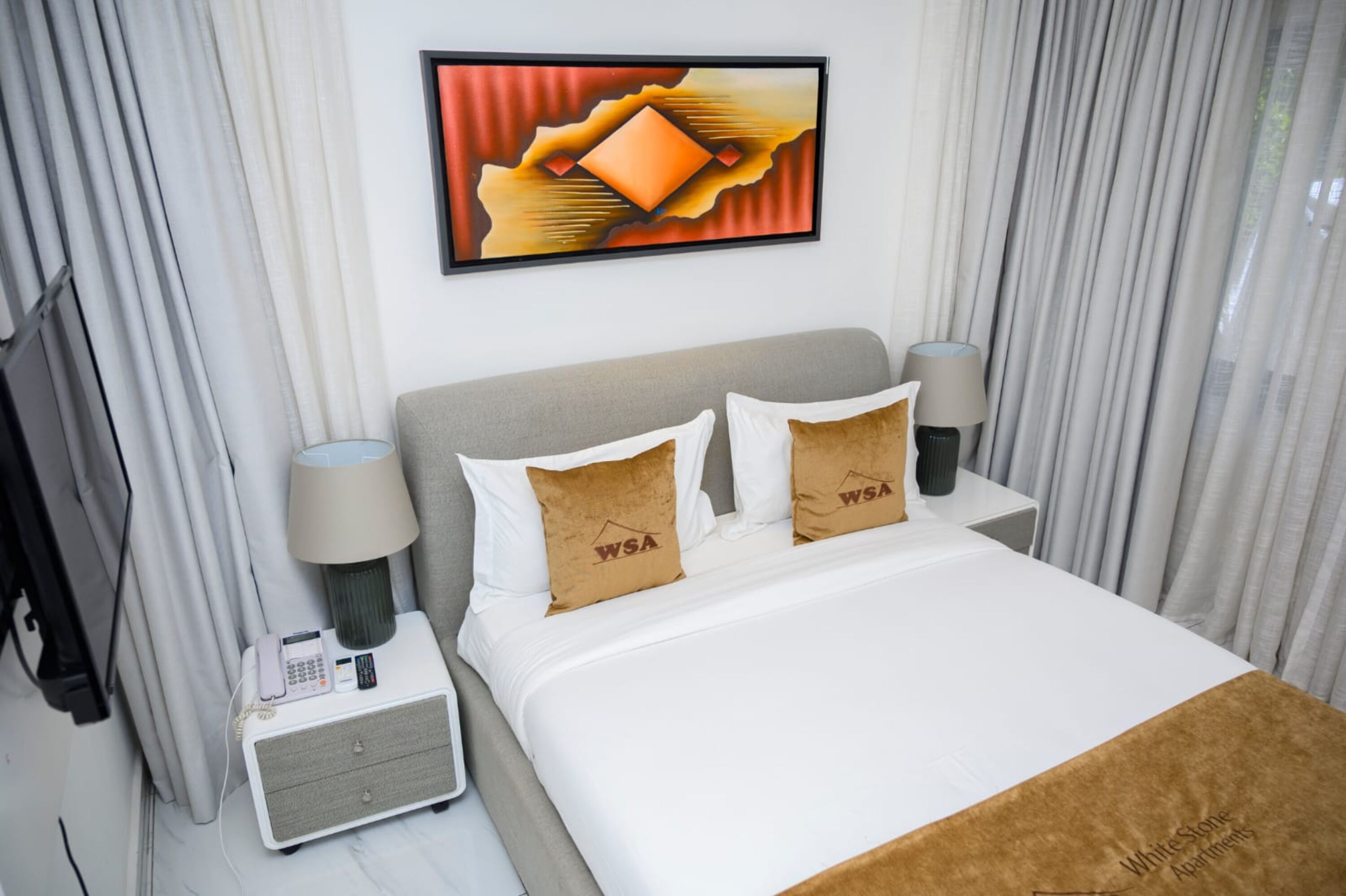 A hotel room with a bed, two pillows with 'WSA' logo, a bedside table with a lamp, a phone, and a painting on the wall.