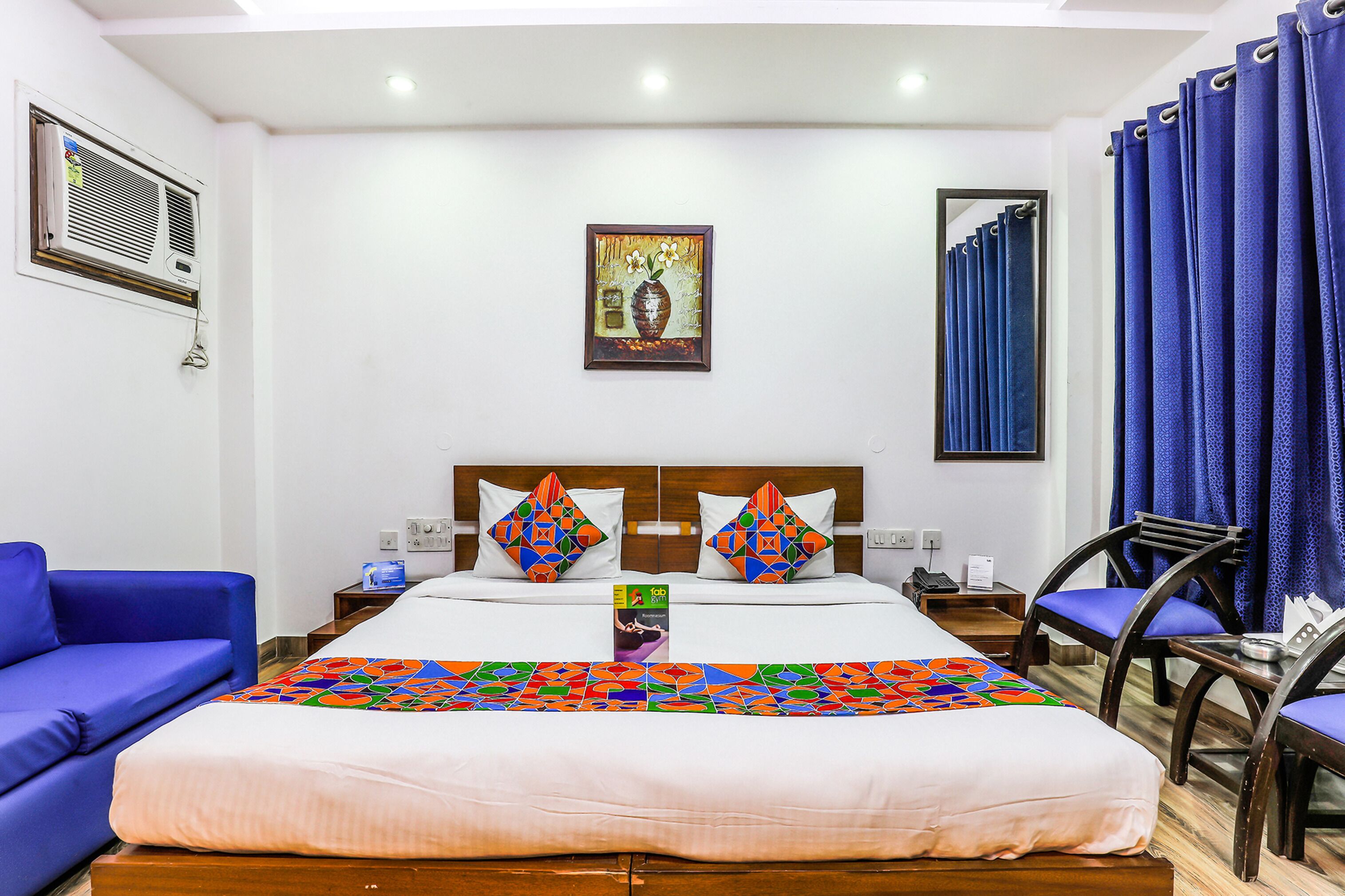 A hotel room with a large bed, two pillows, a nightstand, a chair, a desk, a mirror, a window with curtains, and a painting on the wall.