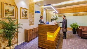 A hotel reception area with a wooden reception desk, a receptionist, and a guest. There are framed pictures on the wall, a sign for 'Fabio Hotels', and potted plants. - Fabhotel Rk International (Mumbai)