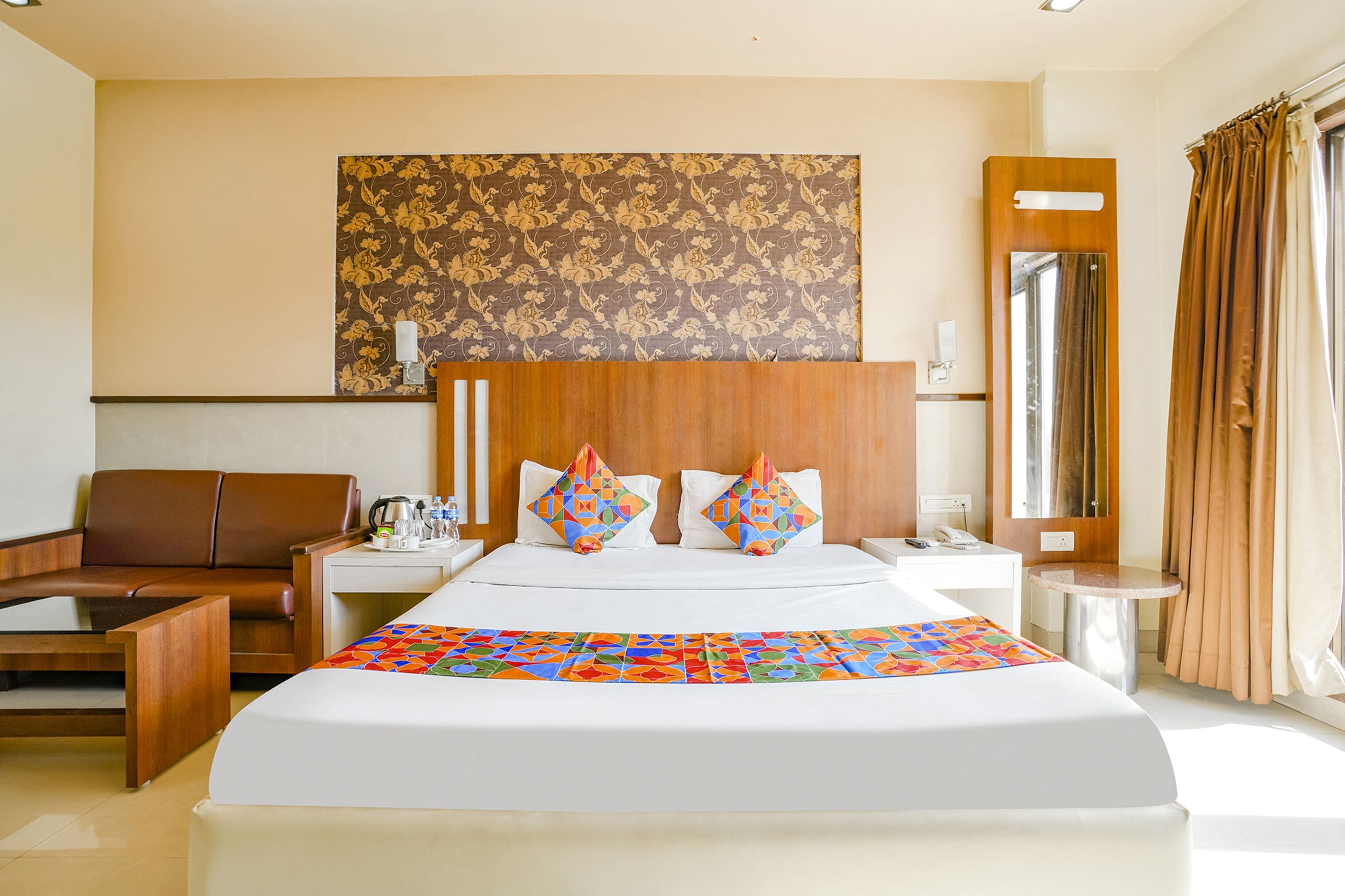 Habitación de hotel con una cama grande, cabecera de madera, mesita de noche, escritorio, silla, espejo y ventana con cortinas.