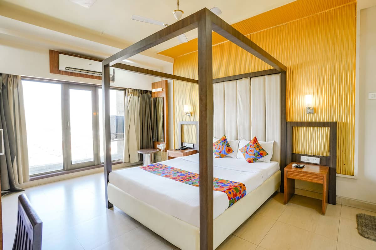 A four-poster bed with a colorful bedspread, a bedside table with a remote control, and a wall-mounted lamp.