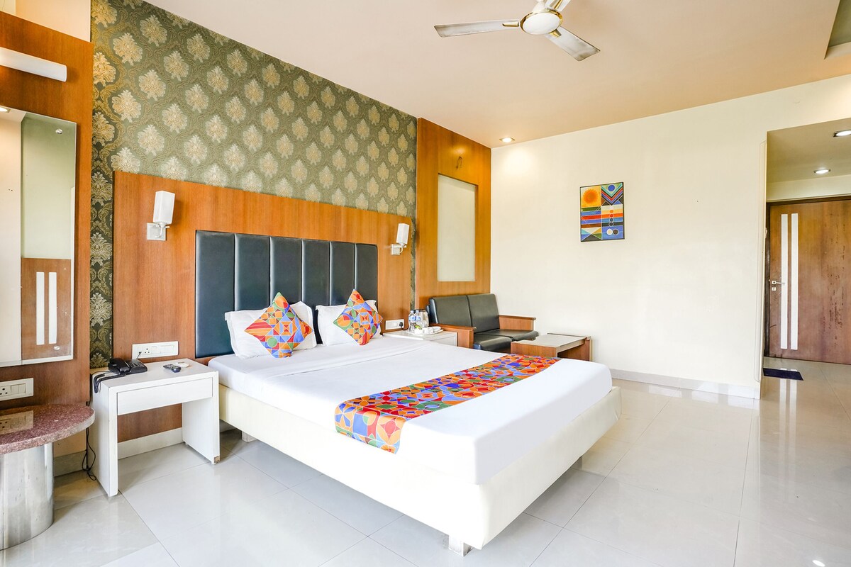 A modern hotel room with a large bed, bedside tables, a desk, a chair, a ceiling fan, and a wall-mounted artwork.