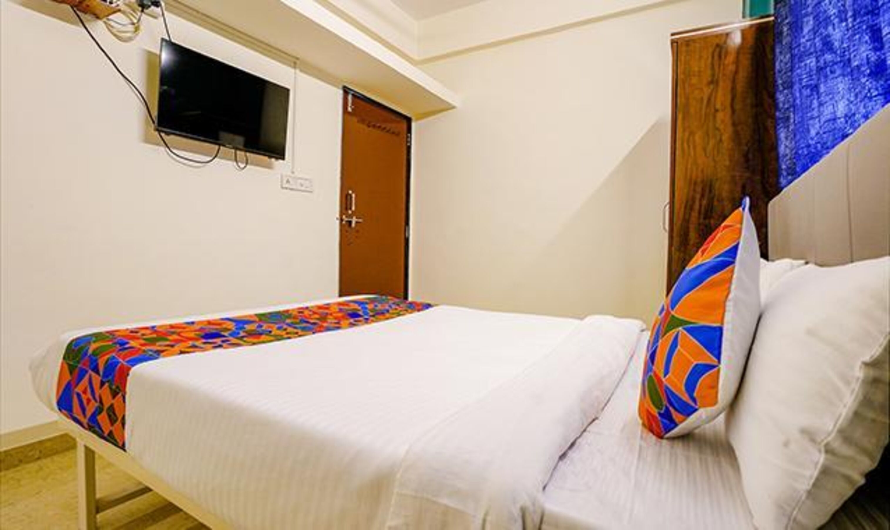 A hotel room with a bed, a television mounted on the wall, and a wooden wardrobe.