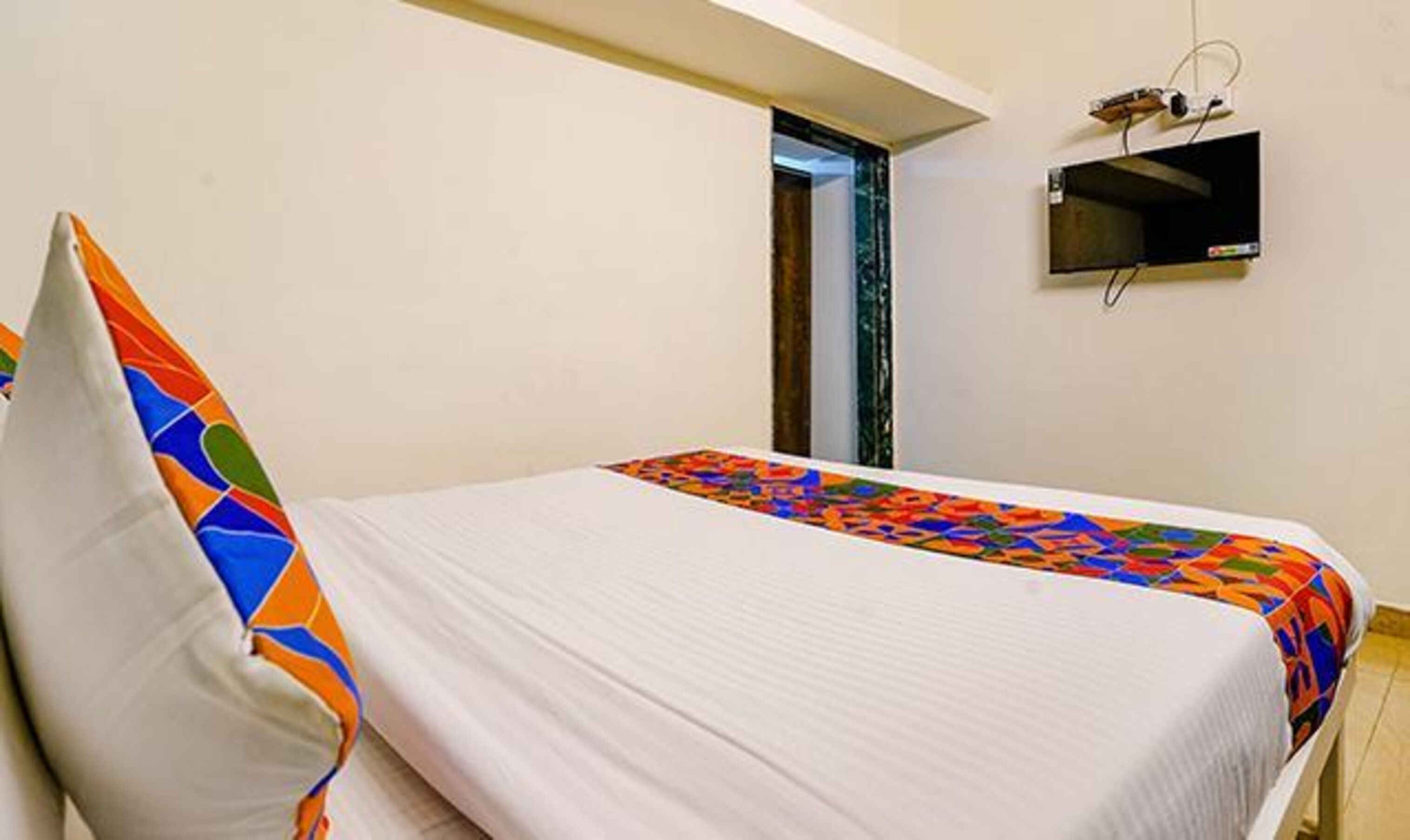 A single bed with a colorful quilt and a white pillow in a room with a flat-screen TV mounted on the wall.