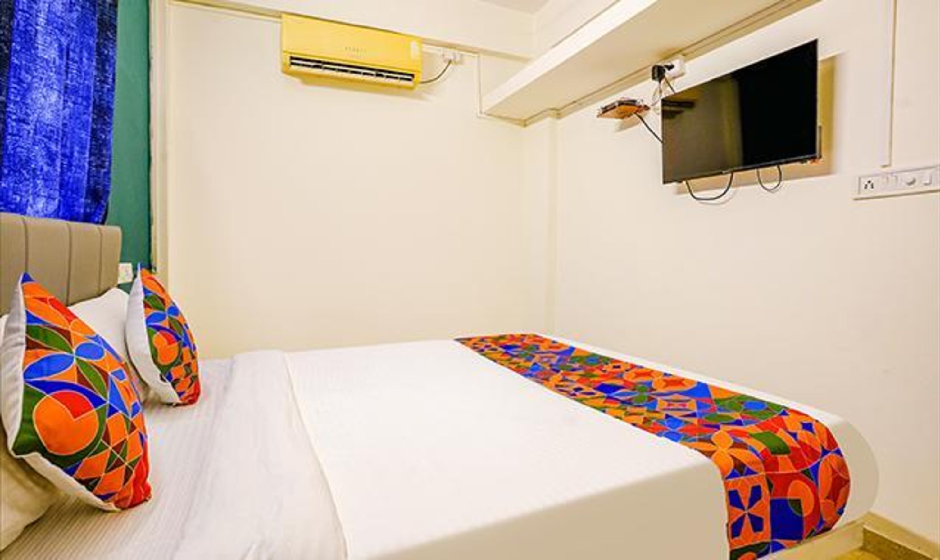 A bedroom with a bed, colorful pillows, a TV, and an air conditioning unit.