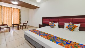 Quarto de hotel com cama, mesa de cabeceira, cadeira, mesa pequena e janela com cortinas.