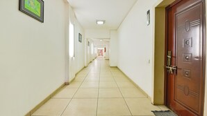Um corredor comprido e estreito com pisos de azulejo e paredes brancas. Quadros emoldurados adornam as paredes e há uma porta de madeira à direita.