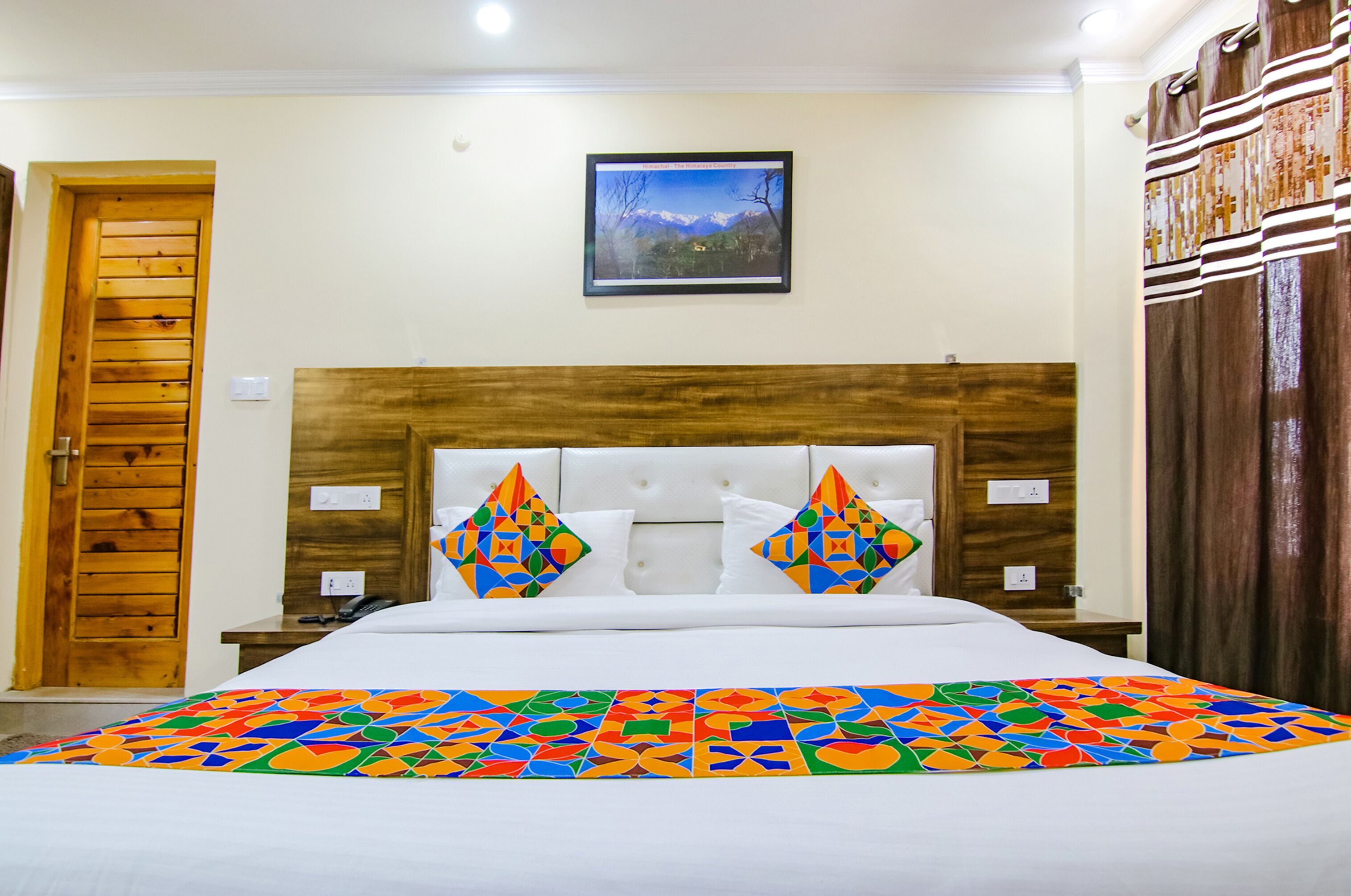 A hotel room with a wooden door, a bed with colorful geometric-patterned pillows, a framed picture on the wall, and a wooden headboard.