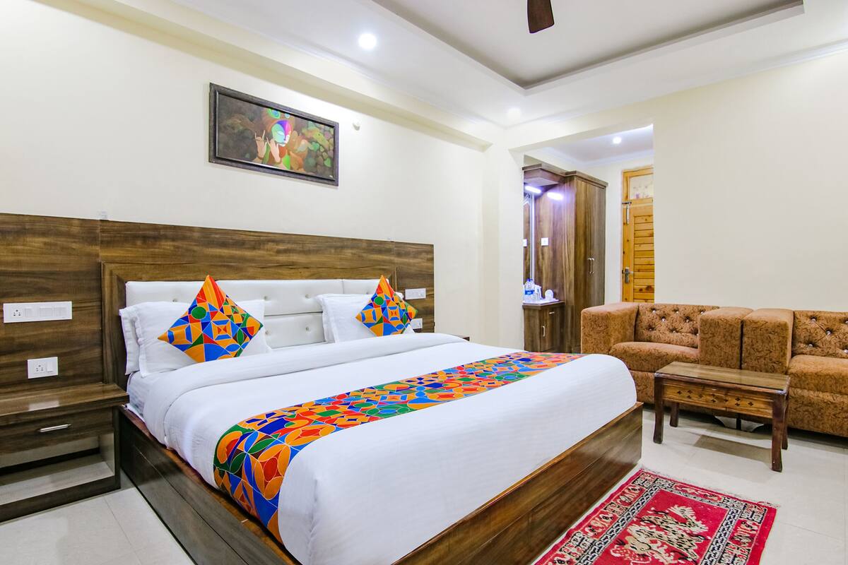 A hotel room with a large bed, a wooden headboard, a colorful bedspread, a red patterned rug, and a brown sofa.