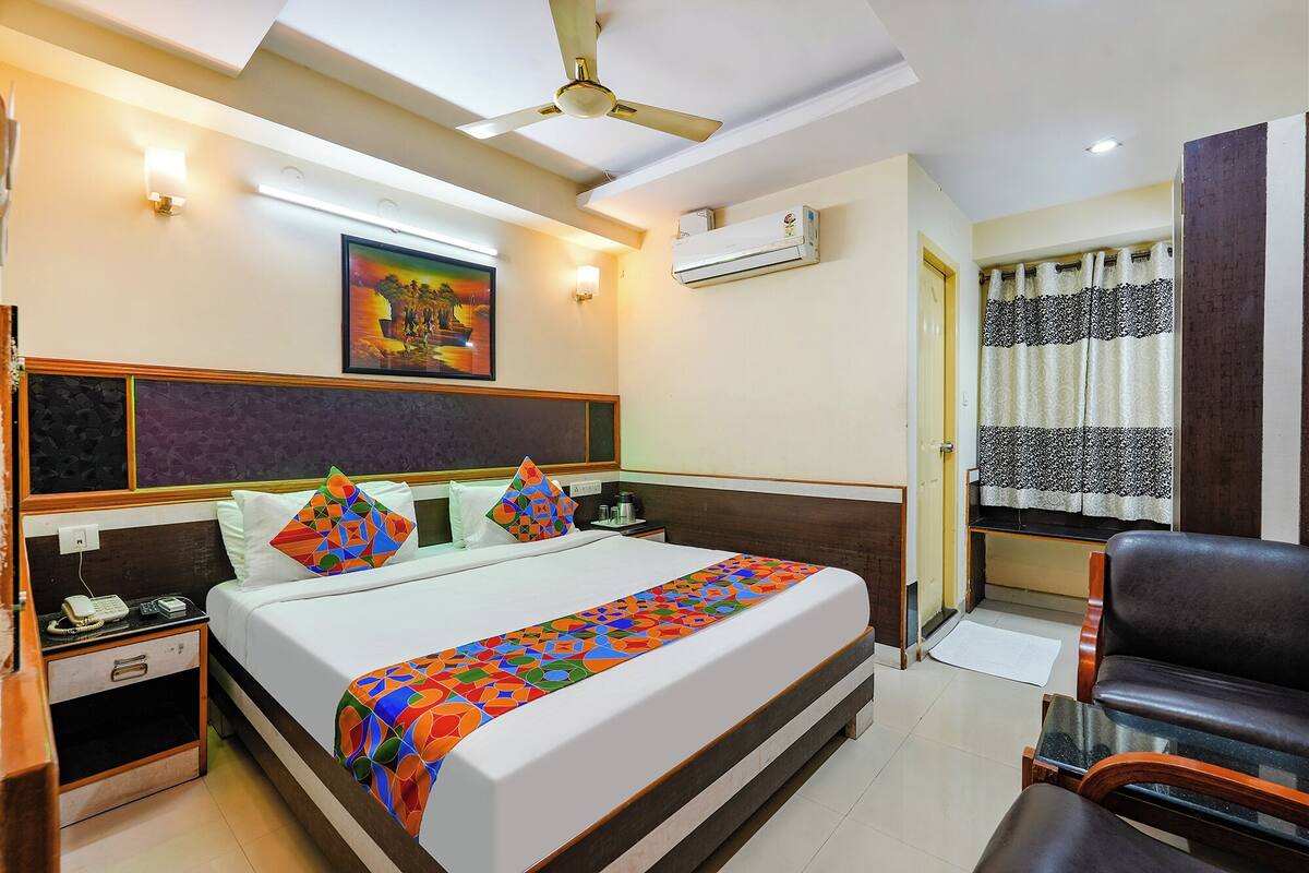 A hotel room with a bed, bedside tables, a chair, a ceiling fan, an air conditioning unit, and a painting on the wall.