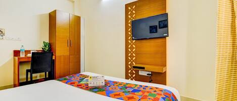 A hotel room with a bed, a desk, a wardrobe, a television mounted on a wooden panel, and a potted plant.