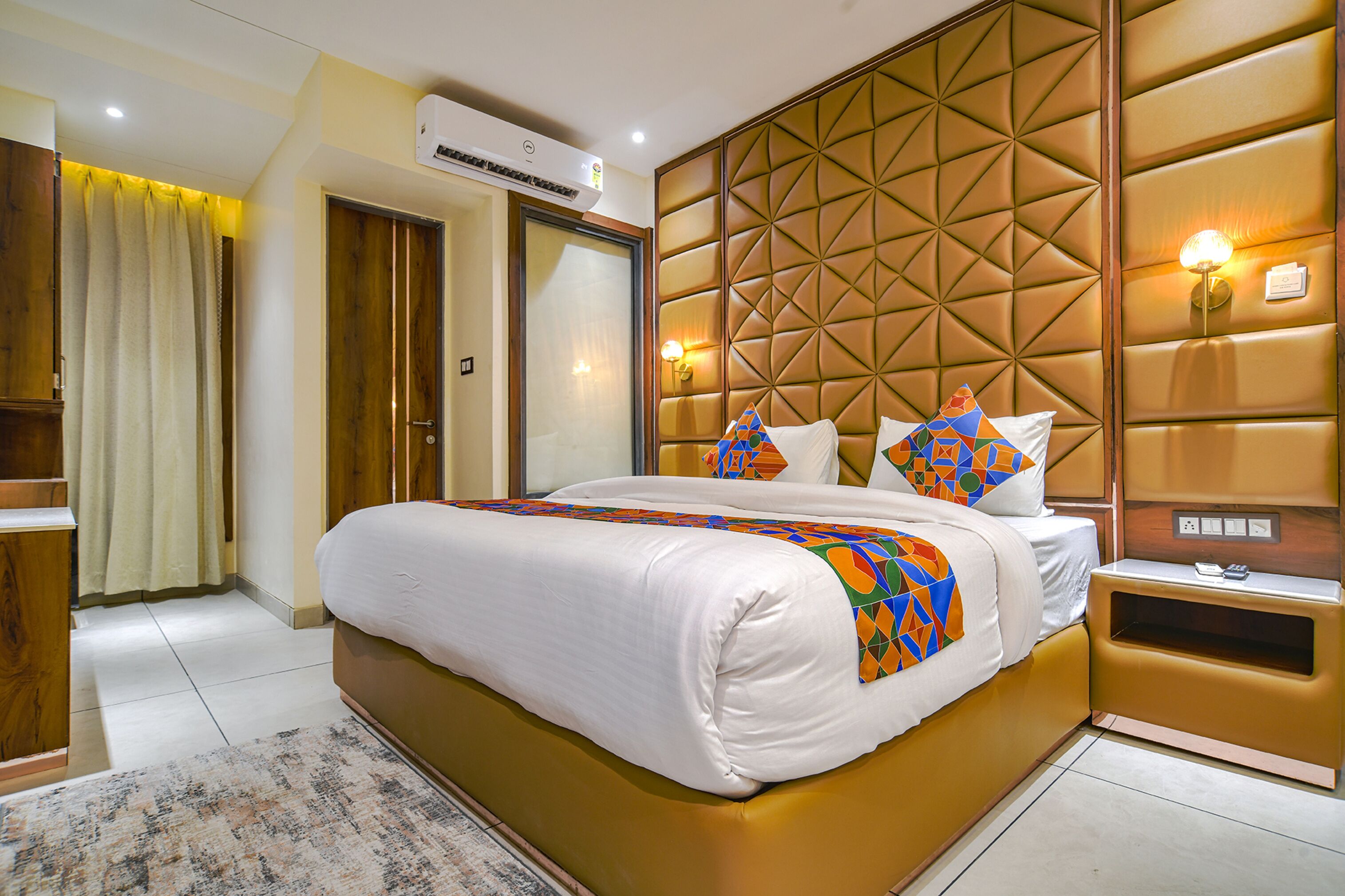 A hotel room with a large bed, a wooden headboard, a nightstand, and a wall-mounted air conditioning unit.