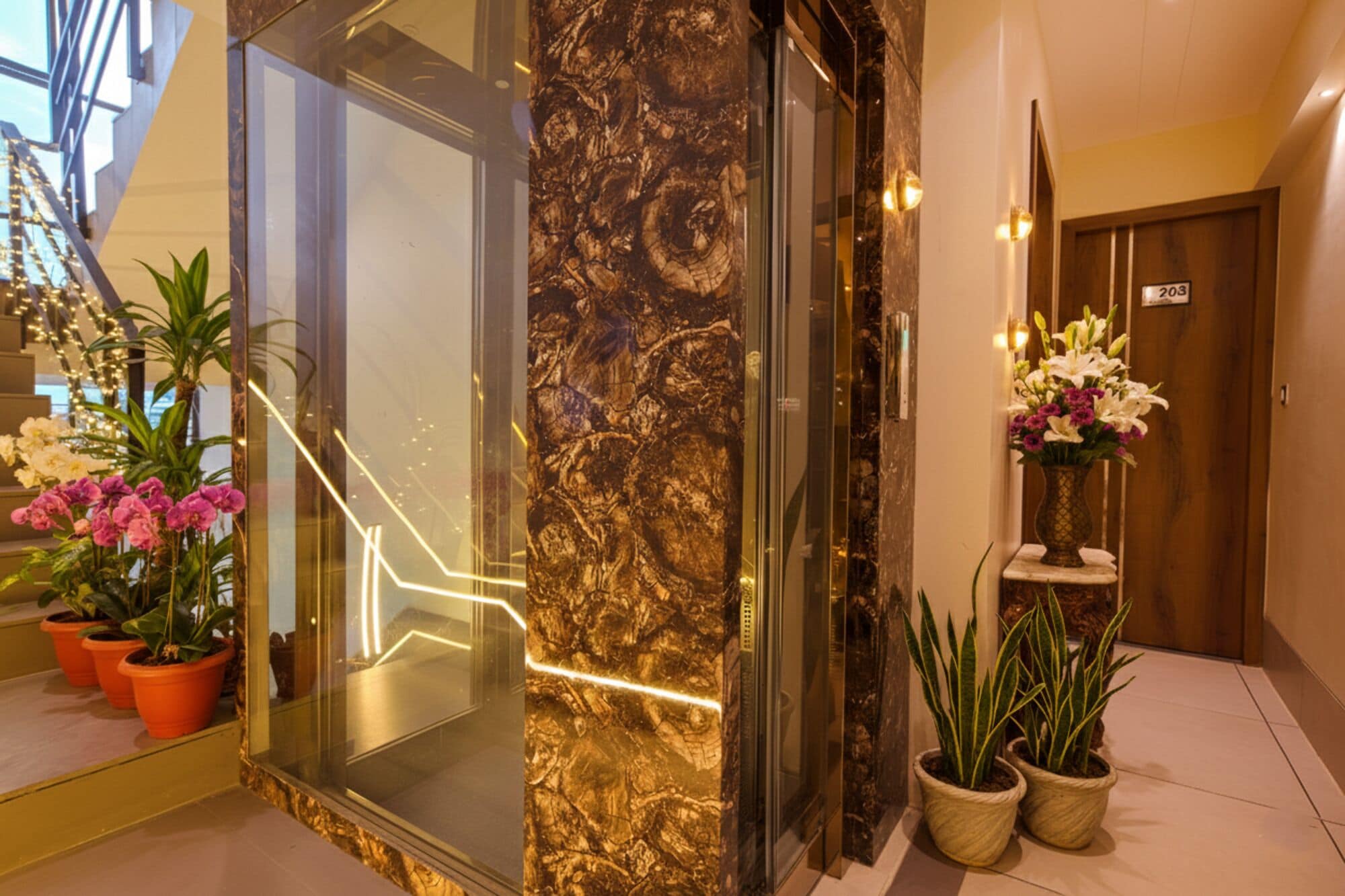 A modern hotel lobby with a glass elevator, marble walls, potted plants, and a wooden door.