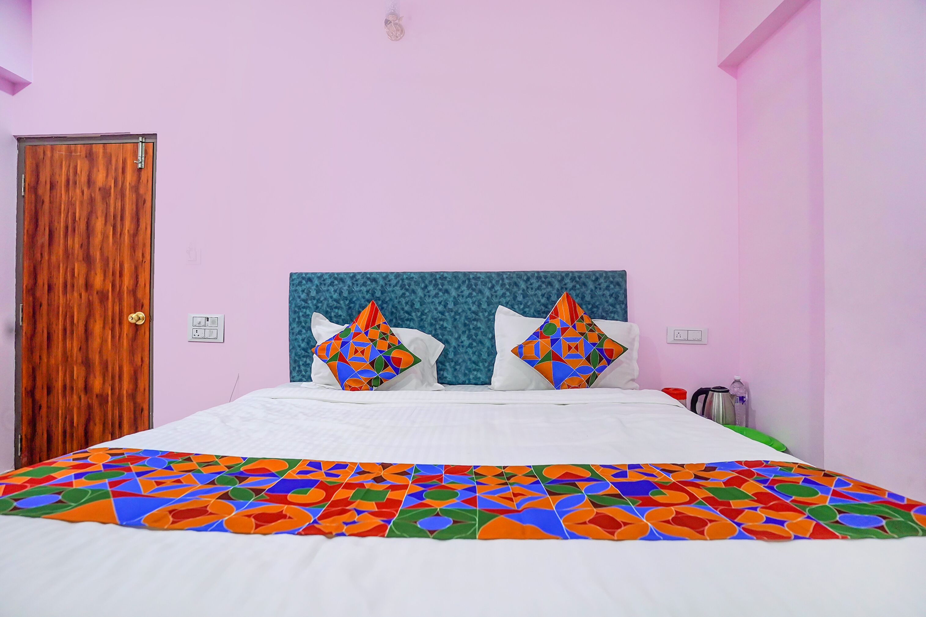 A bedroom with a wooden door, a bed with white bedding, two colorful pillows, and a patterned bedspread.
