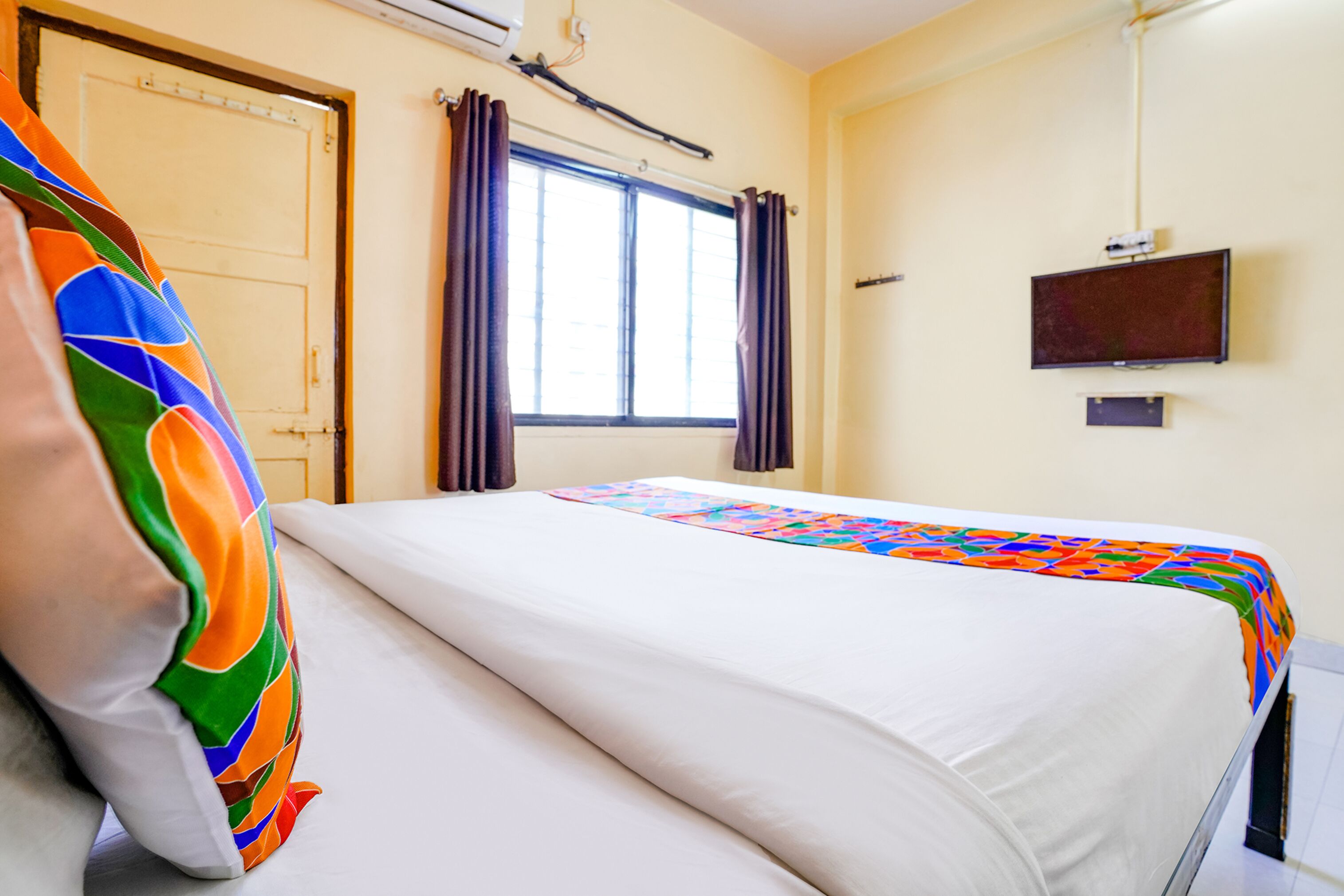 A hotel room with a bed, a window with curtains, a television on the wall, and a door.
