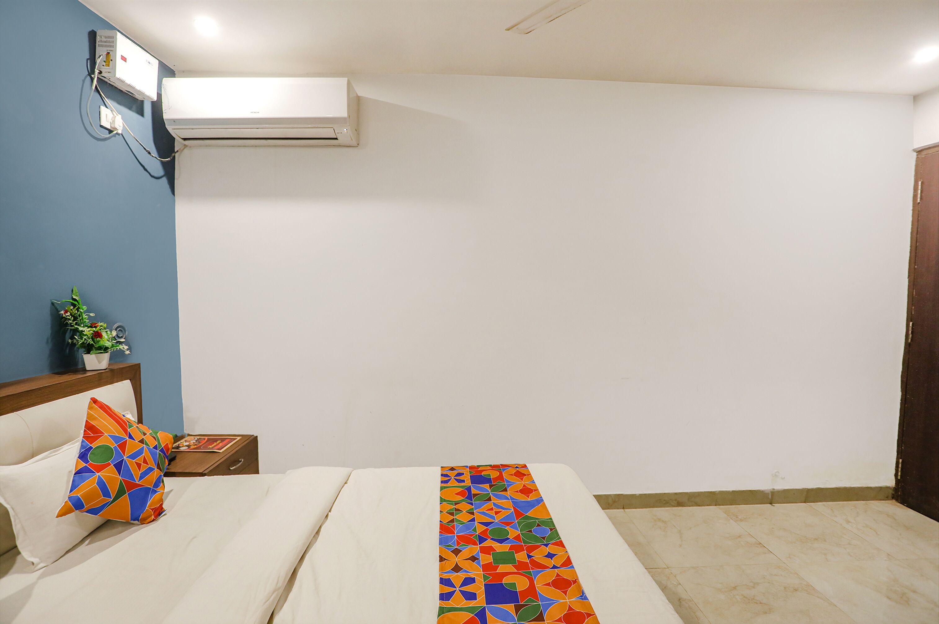 A hotel room with a bed, a nightstand, a potted plant, and a wall-mounted air conditioner.