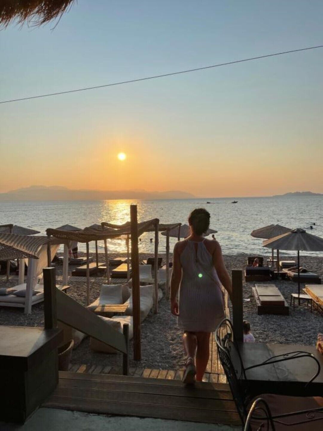 Eine Person geht über eine Holzterrasse zu Strandstühlen und Sonnenschirmen.