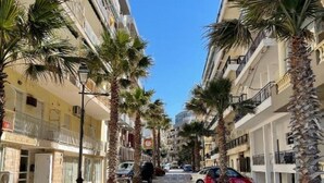 A street lined with palm trees and parked cars, leading to a building with balconies and a clear blue sky. - Sehr Schöne und Moderne Ferienwohnung Lighthouse (Loutraki)
