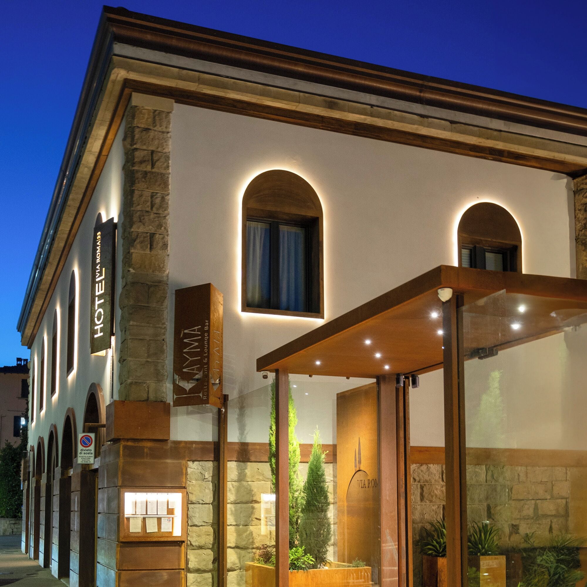 A hotel exterior with a stone facade, arched windows, and a covered entrance area with plants.