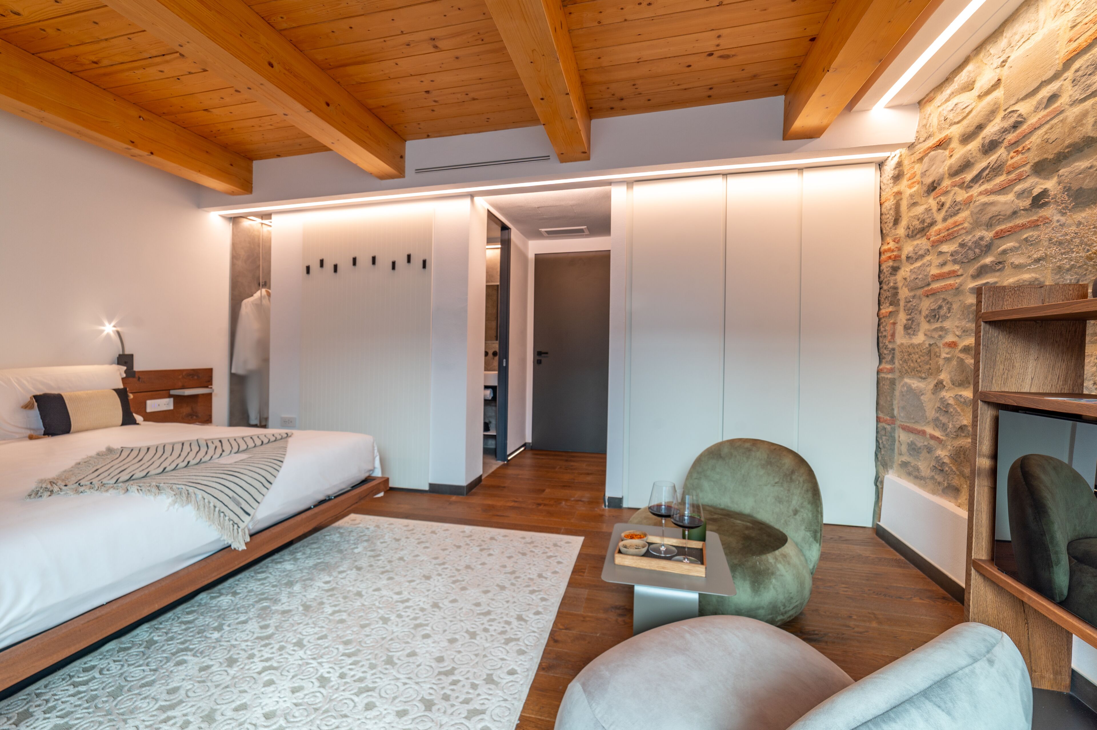 Un dormitorio moderno con techo de madera, una cama grande, pared de piedra y un pequeño rincón de descanso con una mesa.