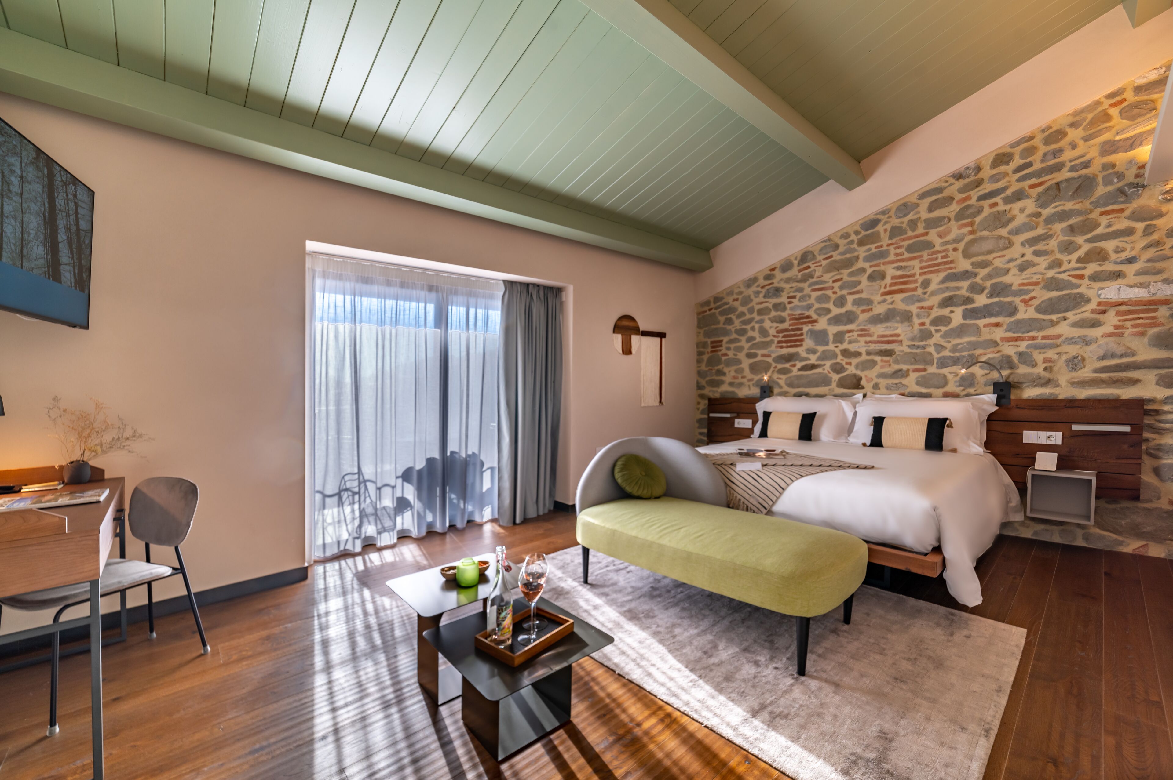 A bedroom with a stone wall, a bed with pillows, a bedside table, a chair, a desk, and a window with curtains.