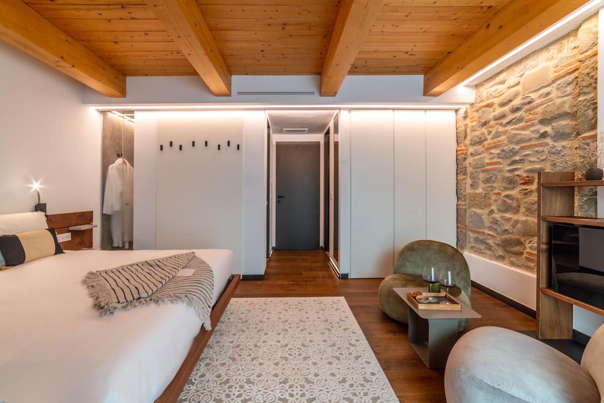 A modern hotel room with a wooden ceiling, a stone accent wall, a bed with a patterned rug, a seating area with a table, and a hallway leading to another room.