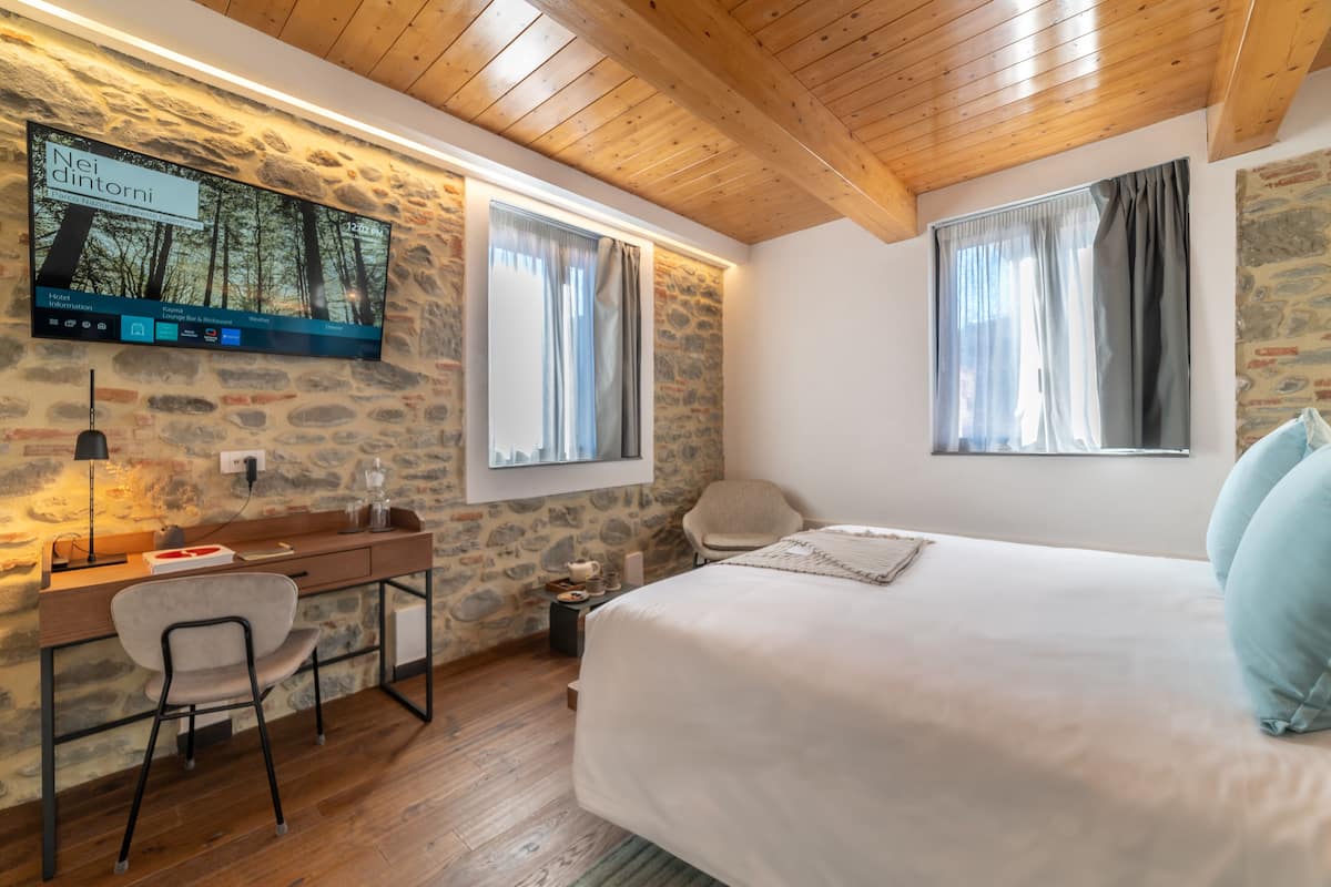 A bedroom with a stone wall, wooden ceiling, a desk with a chair, and a bed with pillows.