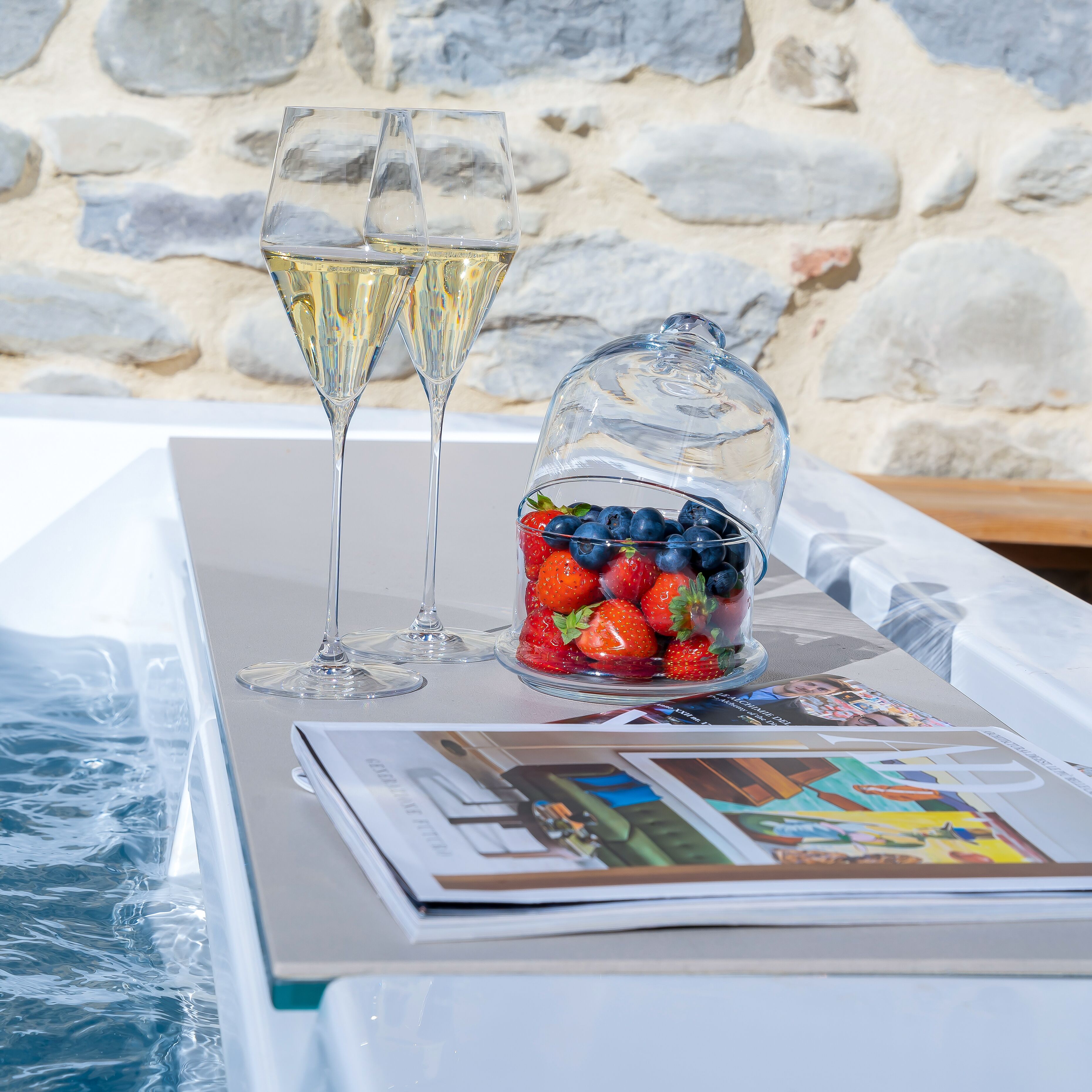 A table with two glasses of white wine, a glass jar of strawberries and blueberries, and a magazine.