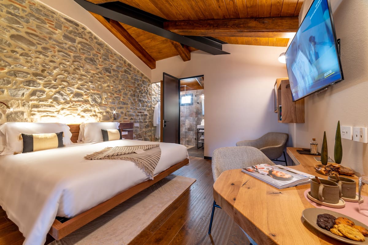 A bedroom with a stone wall, wooden ceiling beams, a bed with white bedding, a TV mounted on the wall, and a breakfast table with pastries and mugs.