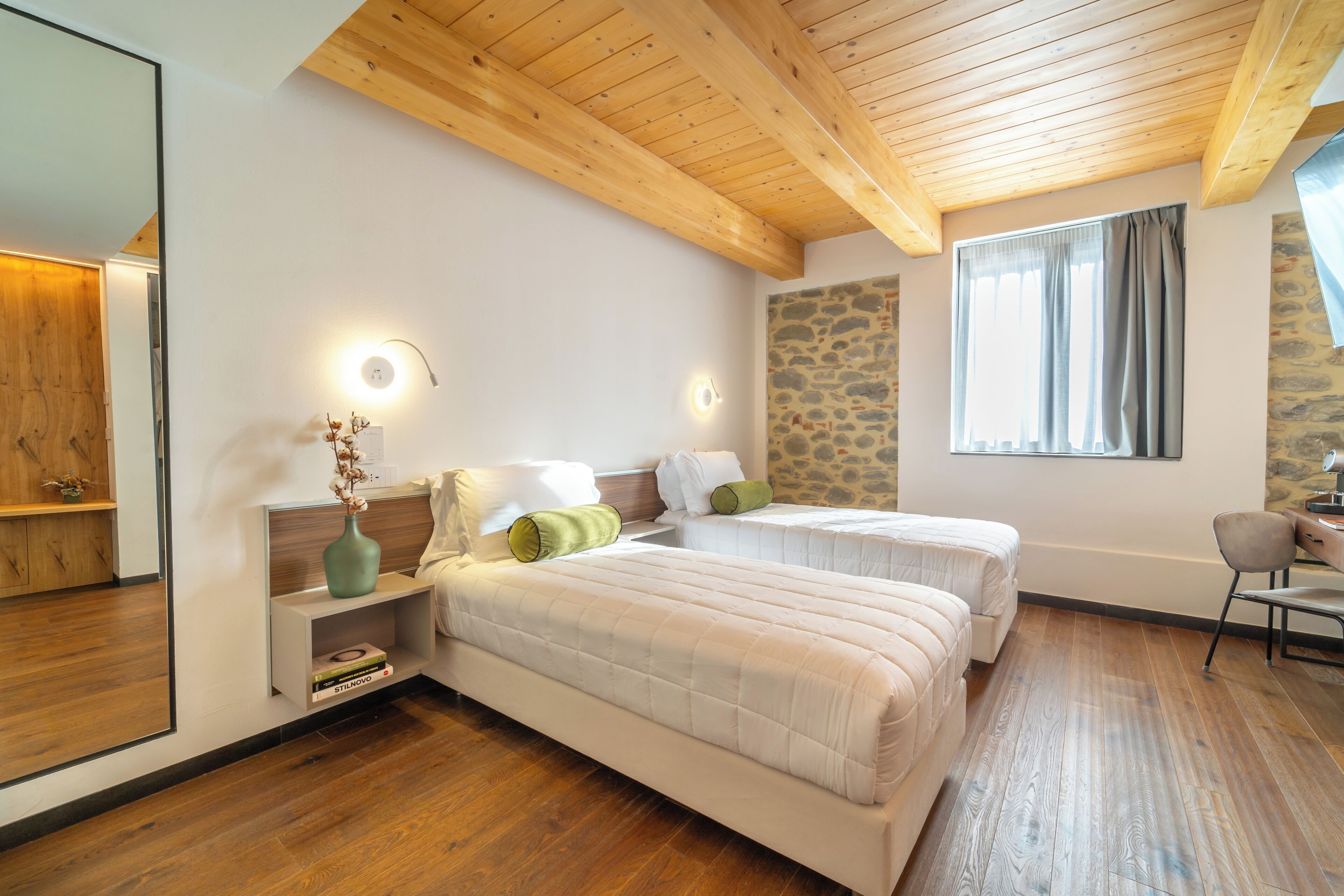 A bedroom with two beds, wooden ceiling beams, a stone wall, a large window with curtains, and a mirror on one wall.