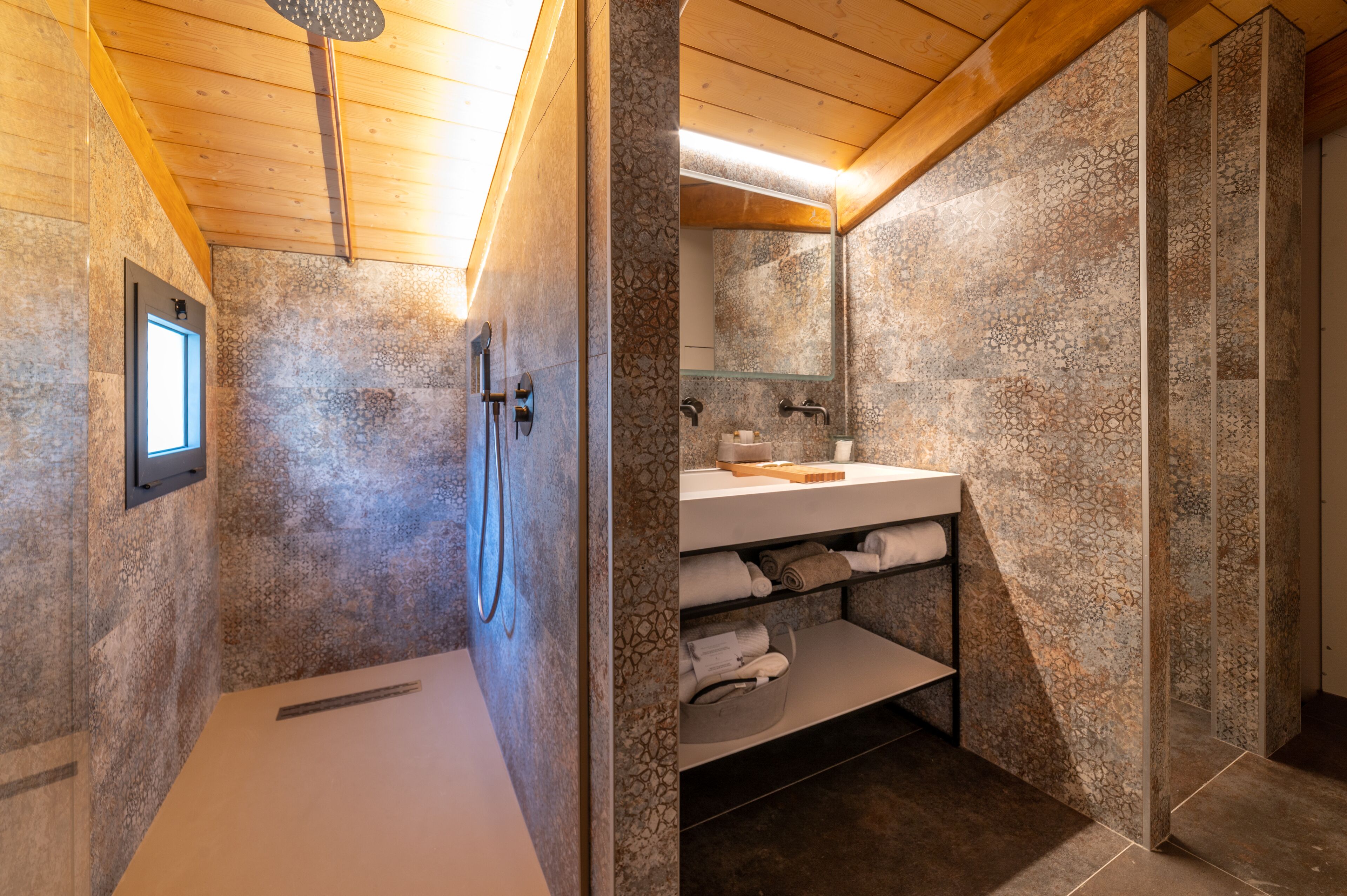 Un baño con paredes de piedra, techo de madera, ducha, lavabo con mampara de vidrio y repisas con toallas.
