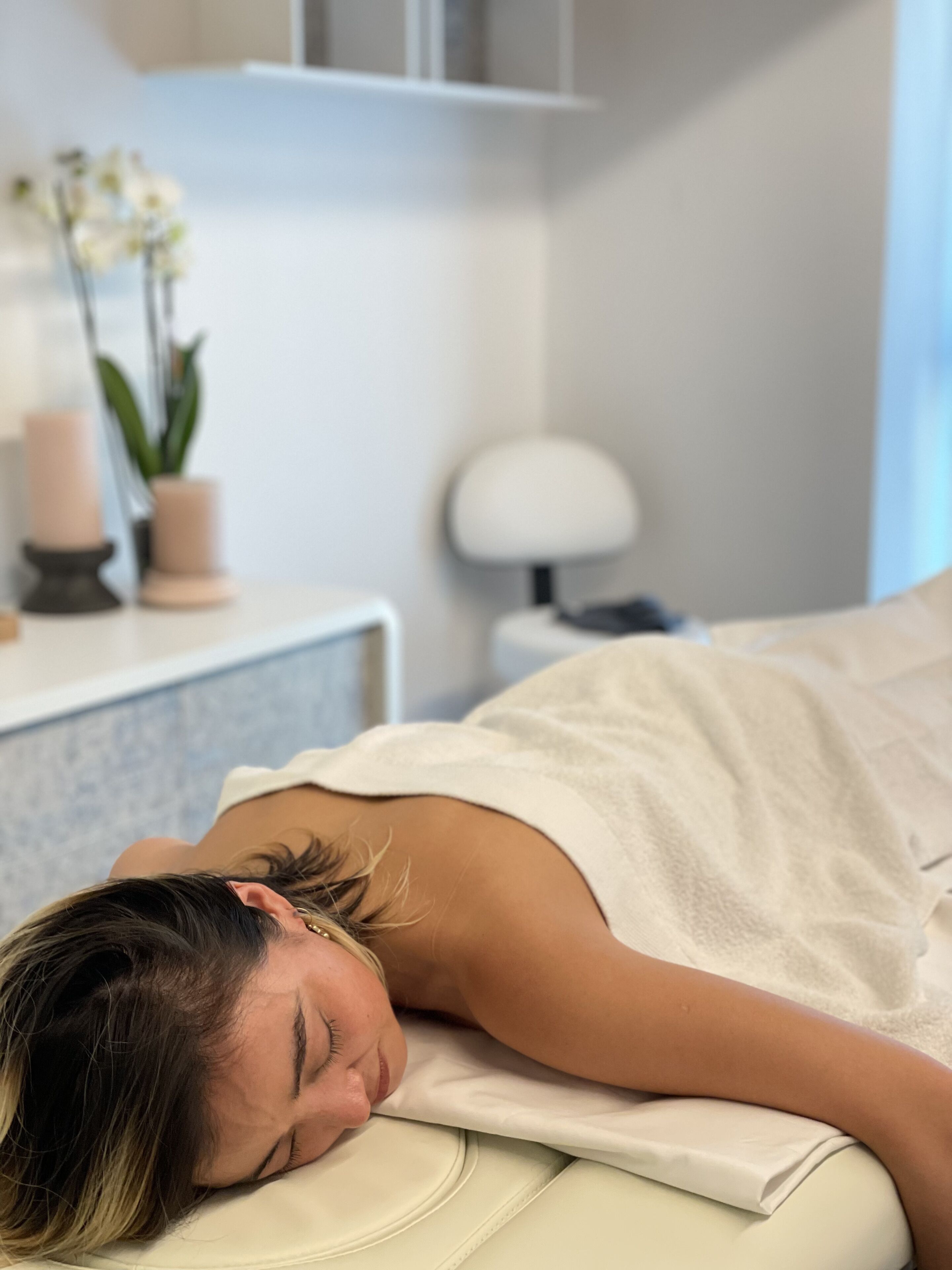A person lying on a bed in a room with a white wall, a small table with a plant, and a lamp.