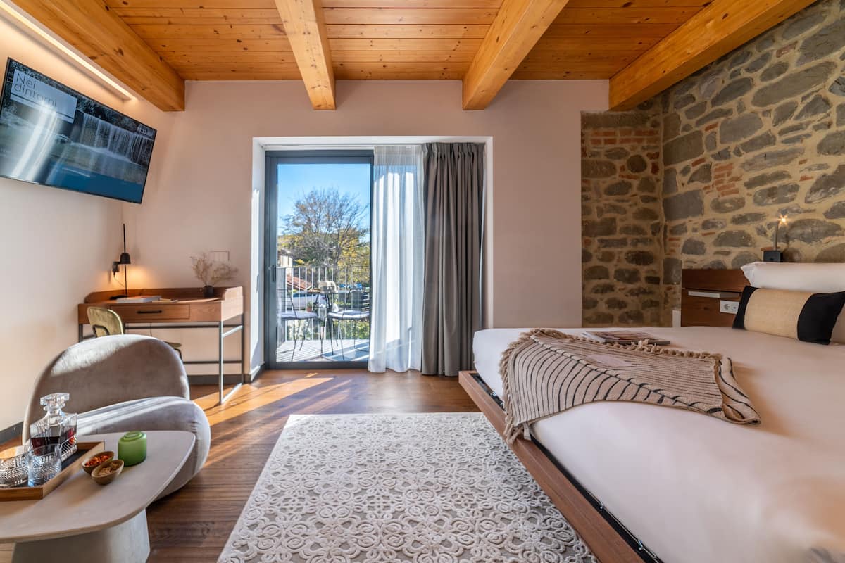 A modern hotel room with a wooden ceiling, a stone wall, a large bed, a desk, and a TV mounted on the wall.