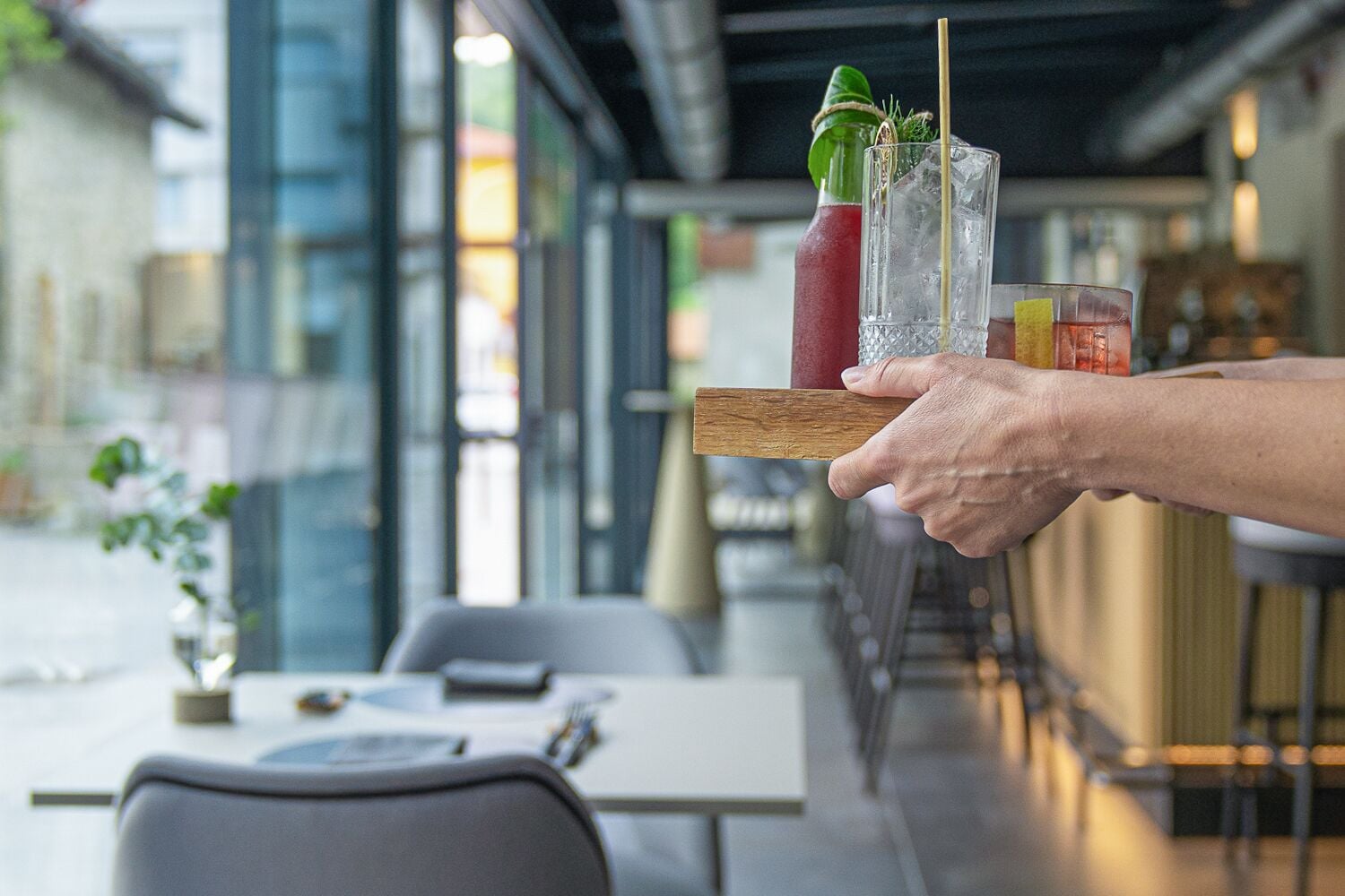 Eine Person hält ein Tablett mit zwei Getränken und einer Flasche in einem modernen Restaurant mit Glaswänden und Tischen.