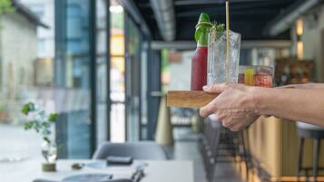Eine Person hält ein Tablett mit zwei Getränken und einer Flasche in einem modernen Restaurant mit Glaswänden und Tischen.