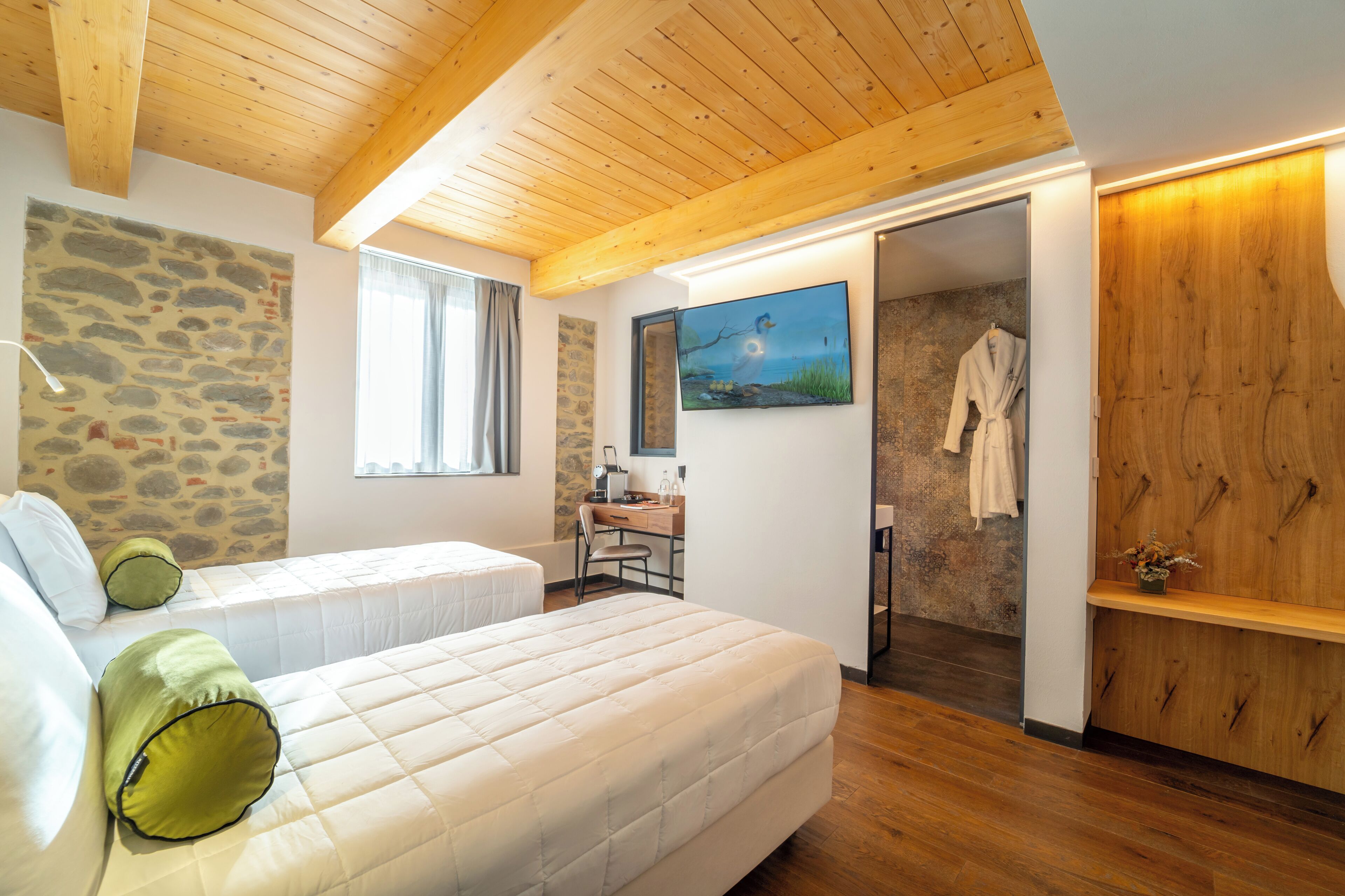 A bedroom with two beds, a stone wall, a wooden ceiling, a desk with a chair, and a bathroom visible through an open door.
