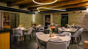 A restaurant with stone walls, wooden ceiling beams, and modern lighting. Tables are set with chairs, and a bar is visible on the left.