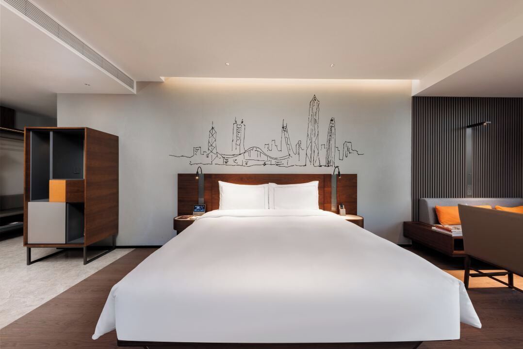 Una habitación de hotel moderna con una cama grande, un cuadro de arte urbano en la pared, una mesita de noche de madera, una mesita auxiliar y una zona de estar con una silla y un sofá.