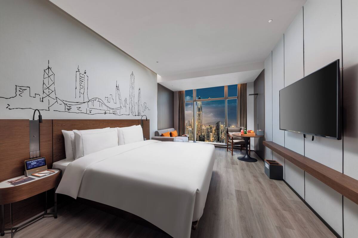 A modern hotel room with a large bed, a flat-screen TV, a desk with a chair, and a cityscape view through the window.