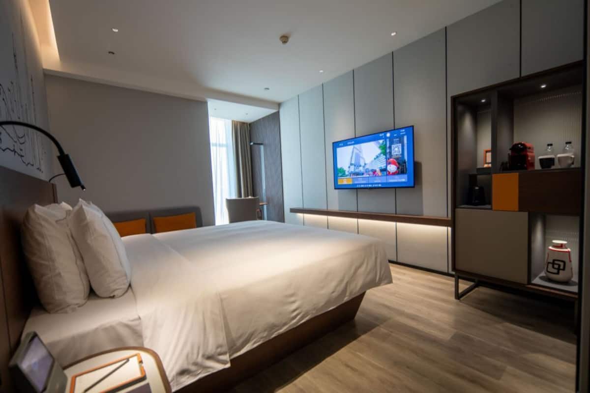 A modern hotel room with a large bed, a flat-screen TV mounted on the wall, and a built-in wardrobe with shelves.