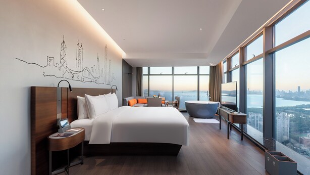 Ein modernes Hotelzimmer mit einem großen Bett, einem Schreibtisch, einem Sessel und Blick auf die Stadt.