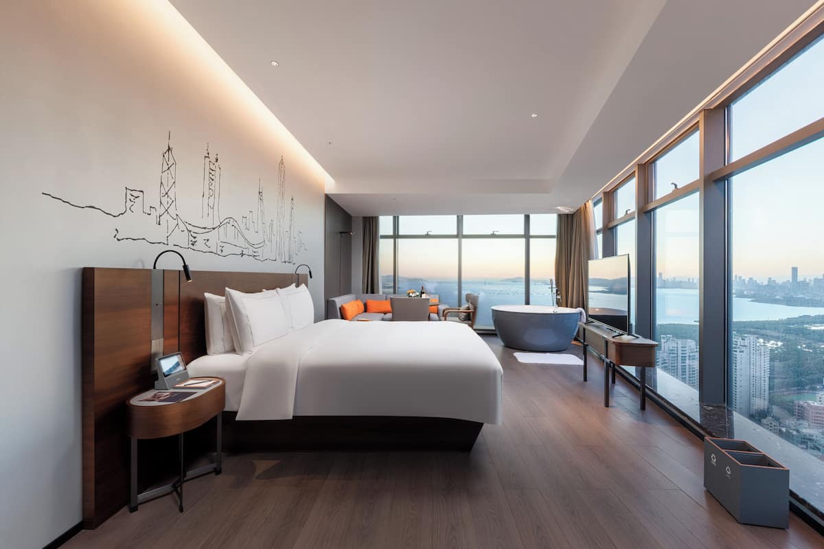 A modern hotel room with a large bed, a desk, a chair, and a view of the cityscape.