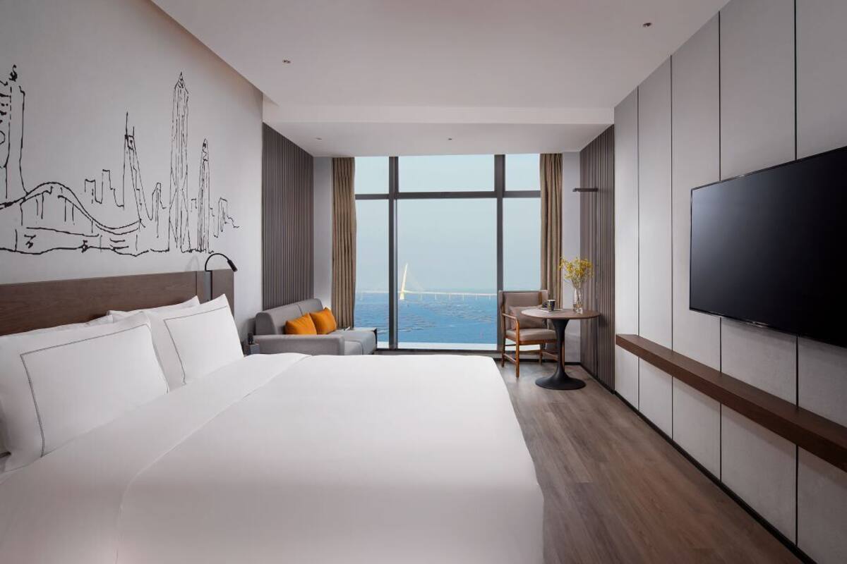 A modern hotel room with a large bed, a TV, a cityscape mural, a view of the sea, and a dining area.