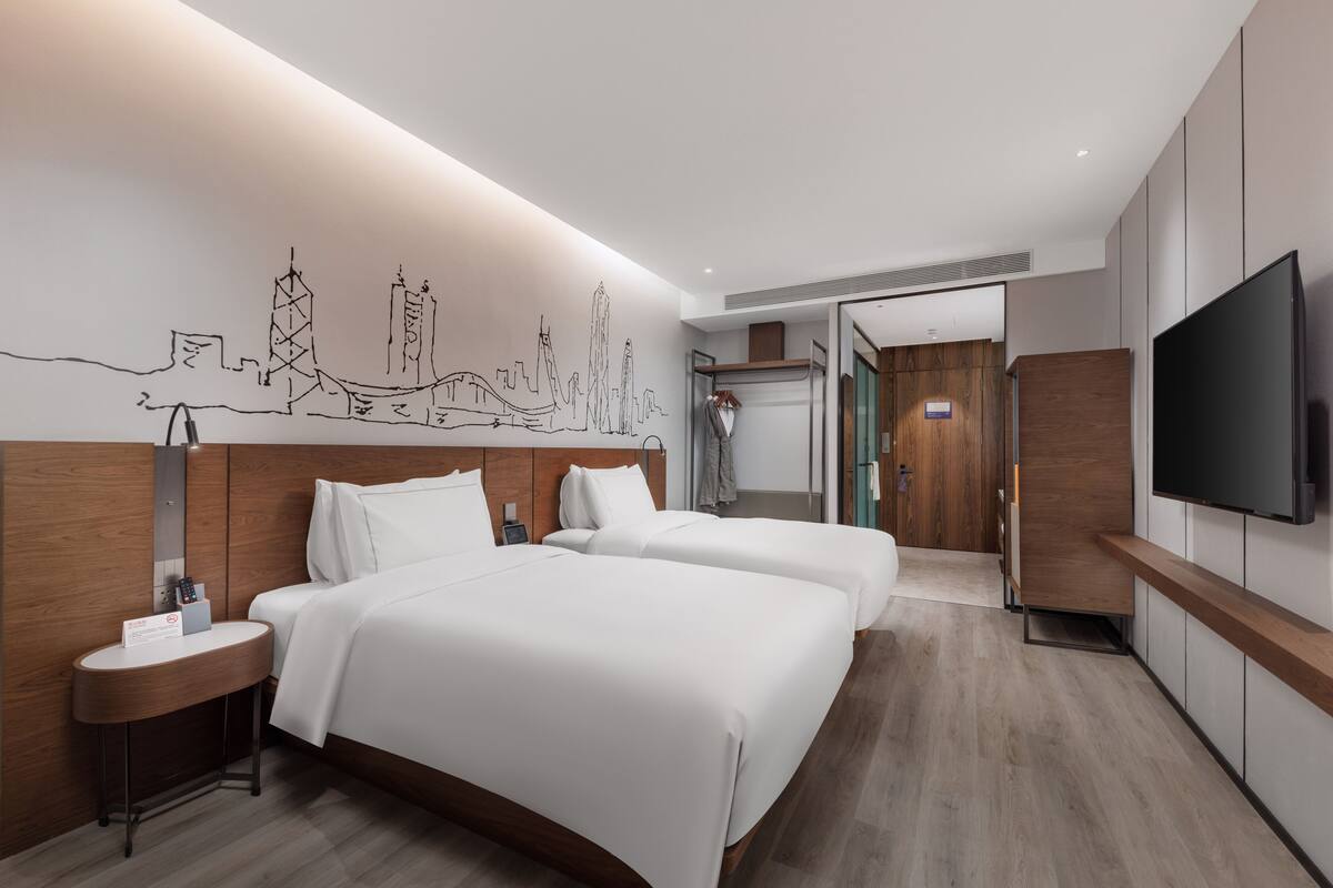A modern hotel room with two beds, a flat-screen TV, a wooden headboard, and a cityscape mural on the wall.