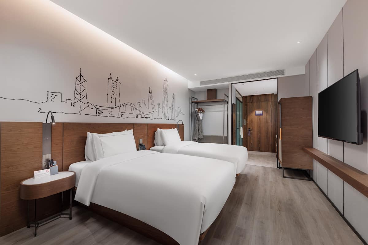 Ein modernes Hotelzimmer mit zwei Betten, einem Flachbildfernseher, einem hölzernen Kopfteil und einem Wandgemälde mit Stadtansicht.