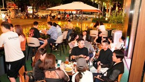 Eine Gruppe von Menschen speist draußen in einem Restaurant mit Tischen und Stühlen, umgeben von Pflanzen und Straßenlaternen.