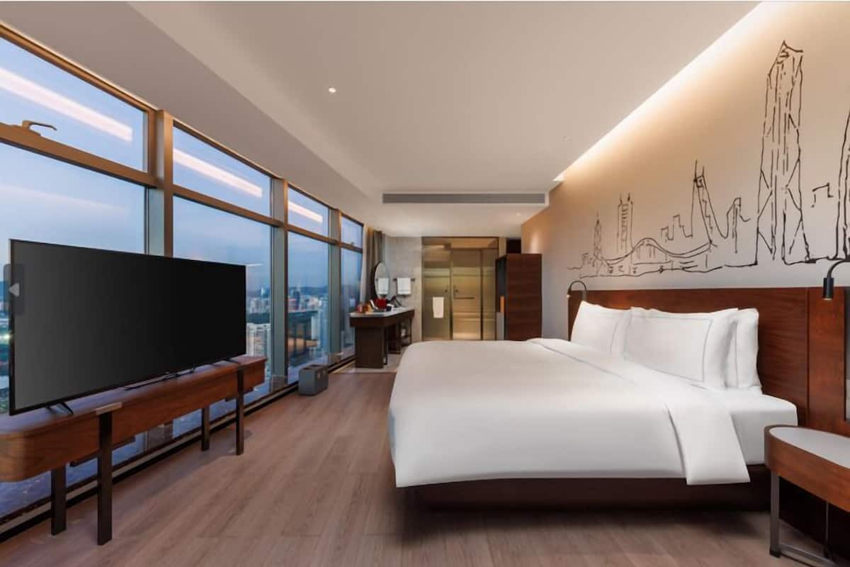 A modern hotel room with a large bed, a flat-screen TV on a wooden stand, a city view through large windows, and a wall mural.